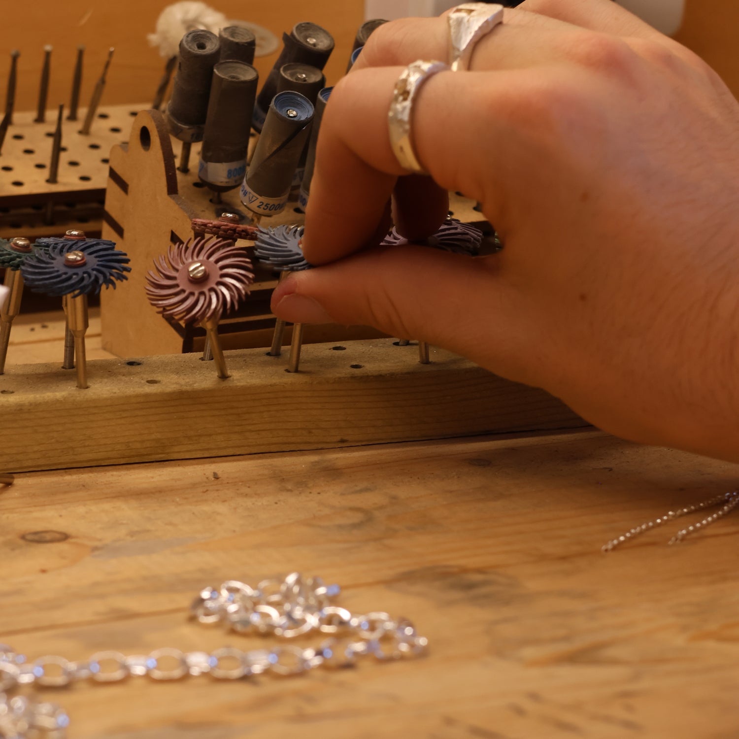 A hand selects a rotary tool attachment from a wooden holder on a workbench, with metal chains and various tools visible.