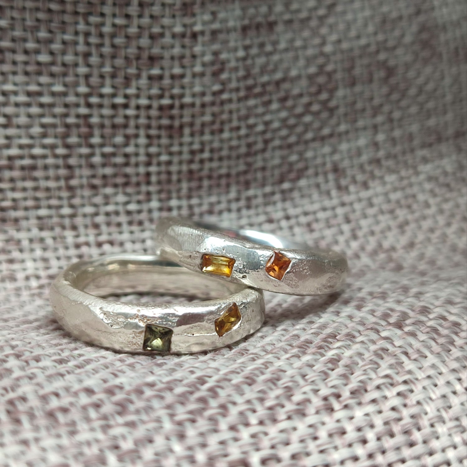 Two silver rings with rough-textured bands, each set with two small rectangular gemstones in green and amber-orange hues, displayed on a woven fabric background.