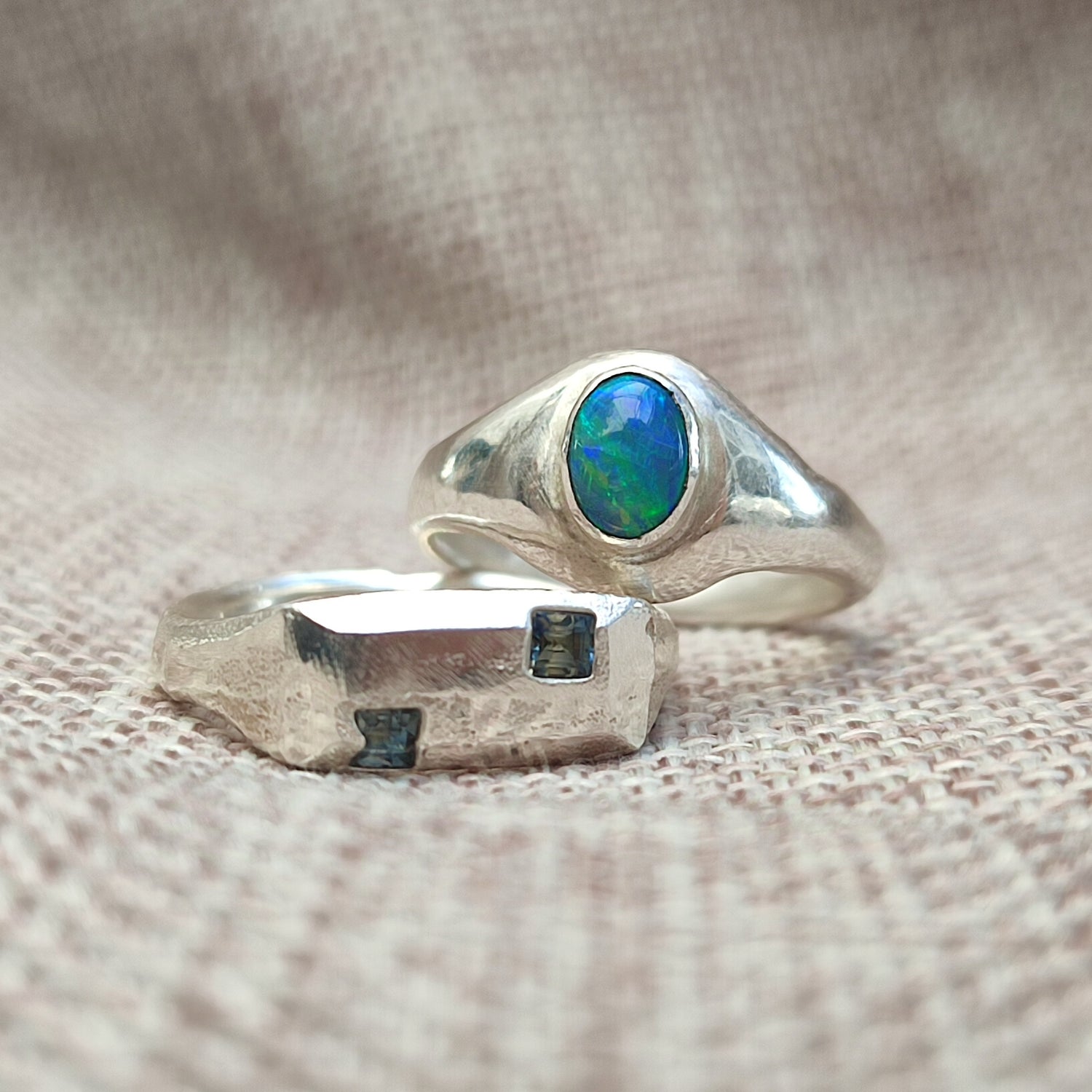 Two silver rings on textured fabric; one features an oval blue-green opal, the other has a rough finish with two small rectangular blue stones.
