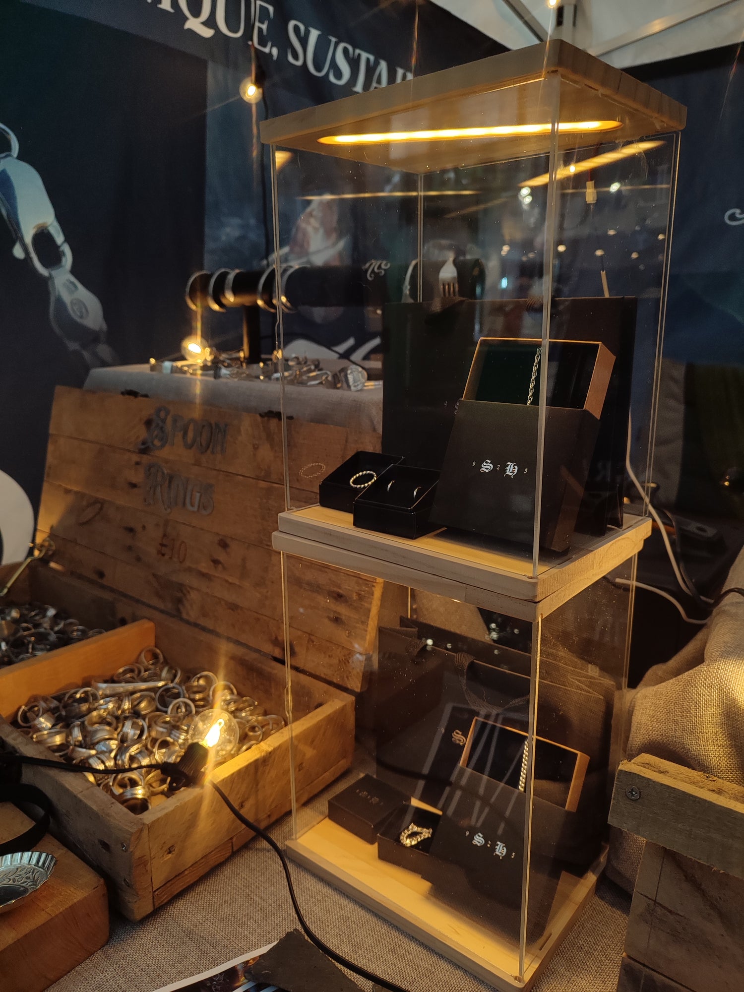 A display case with illuminated shelves showcases rings and jewelry; a wooden box with metal rings and a sign reading "Spoon Rings £10" sits beside it.