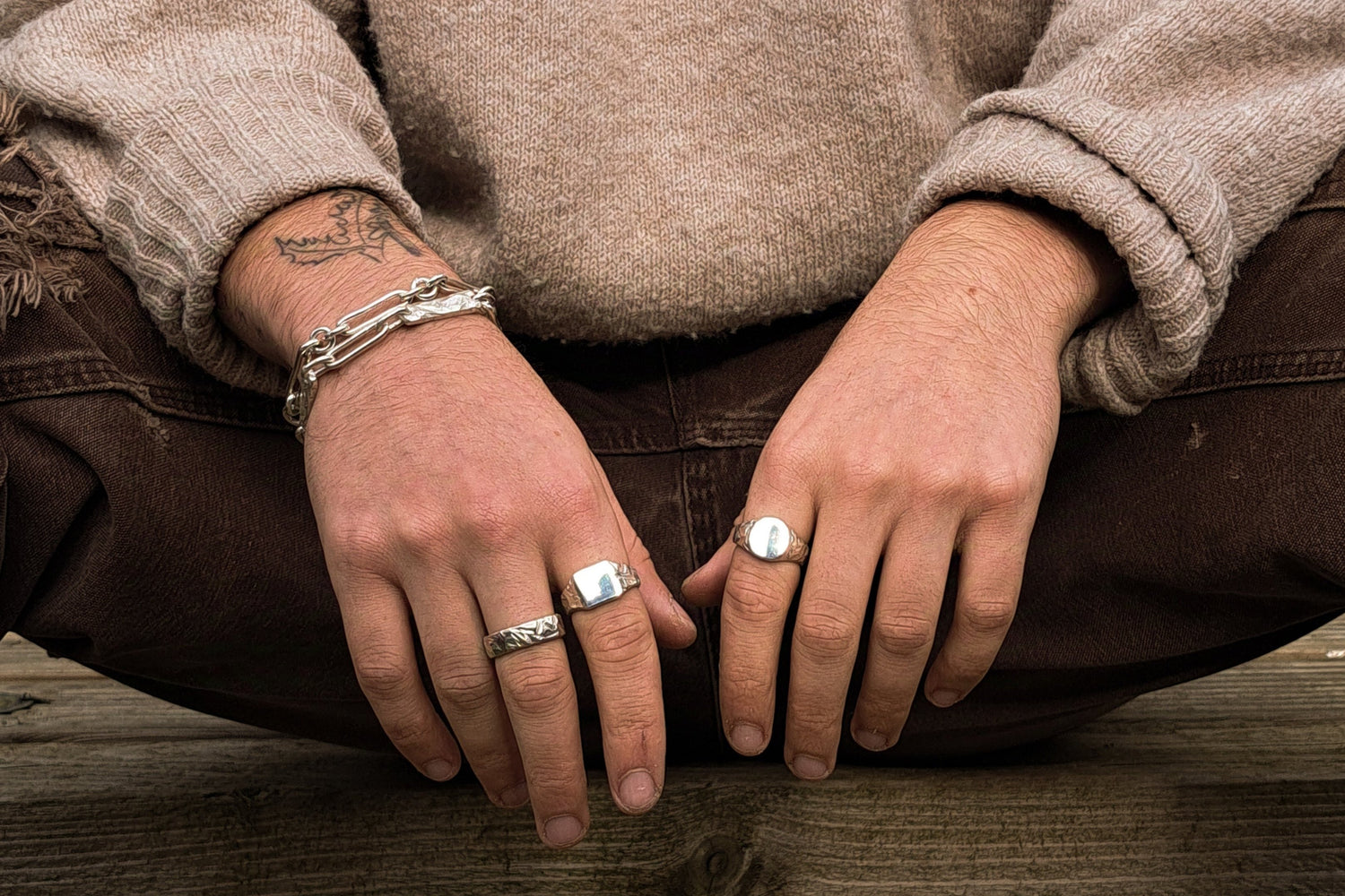 A person sits cross-legged, wearing a beige sweater, brown pants, silver rings on both hands, a chain bracelet, and has a visible forearm tattoo.