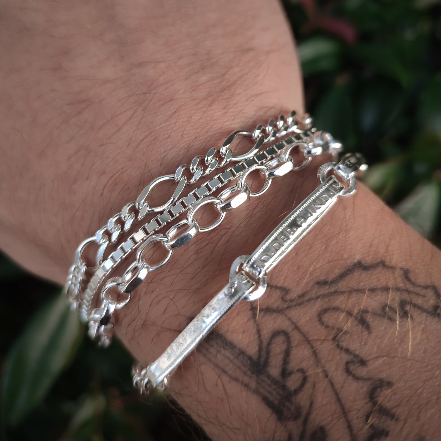 A close-up shows a person's wrist wearing three sterling silver chain bracelets, including the silverhollowjewellery Box Chain Bracelet, with a partial tattoo visible and leafy background.