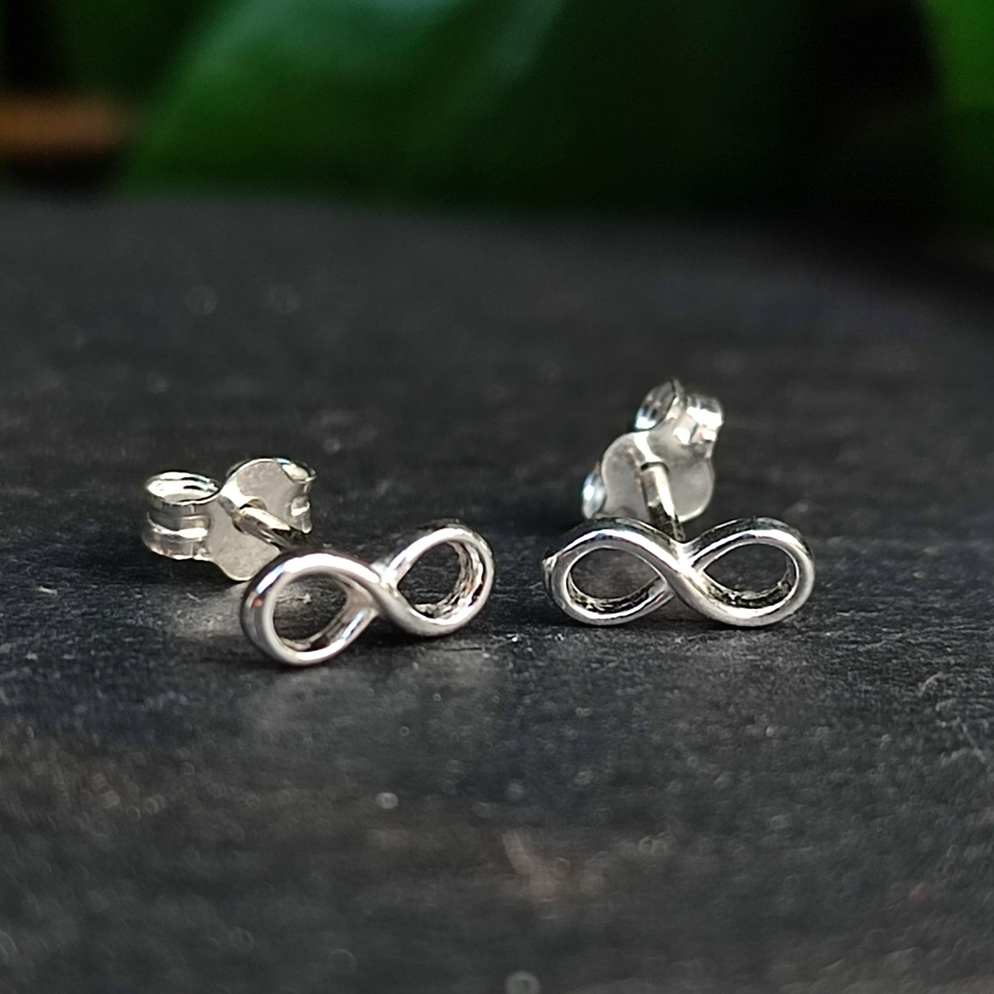 Infinity Stud Earrings-Sterling Silver by silverhollowjewellery: 925 sterling silver studs shaped like elegant infinity symbols, displayed on a dark surface with a blurred green background.