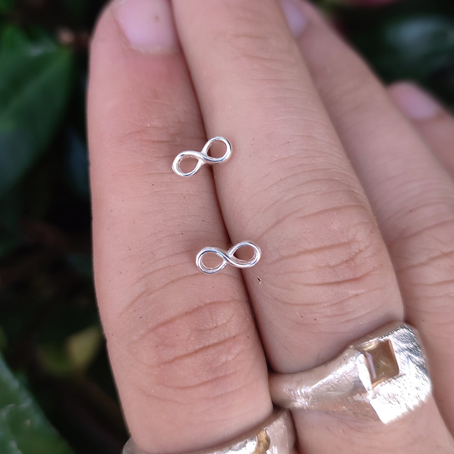 Two fingers wearing silverhollowjewellery’s Infinity Stud Earrings—Sterling Silver and a silver ring with a square gem, set against a blurred leafy background.