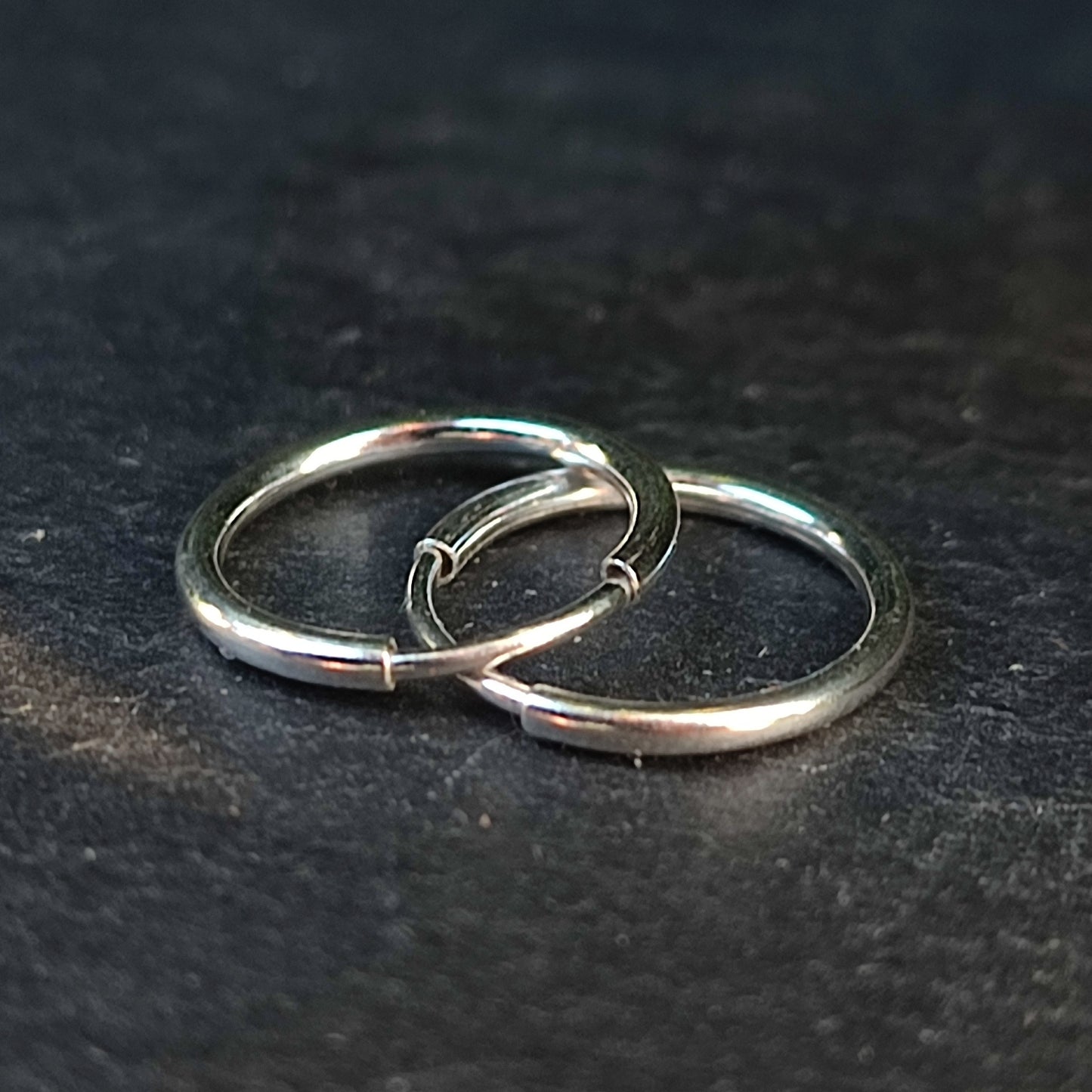 Two silverhollowjewellery Sterling Silver 12mm Hoop Earrings, finely polished, are lying flat and interlocked on a dark textured surface.