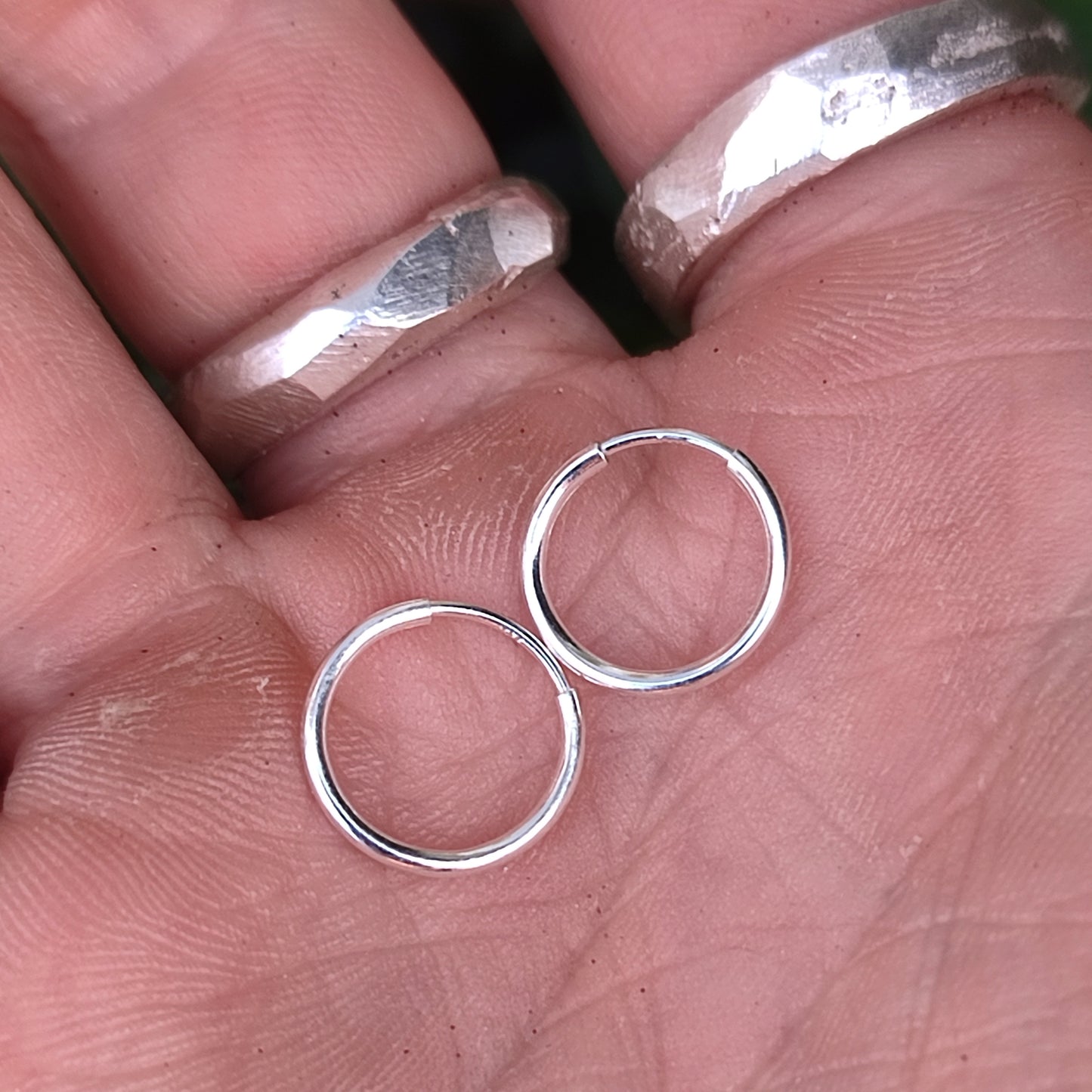 A hand with silver rings holds two small, polished Sterling Silver 12mm Hoop Earrings by silverhollowjewellery.