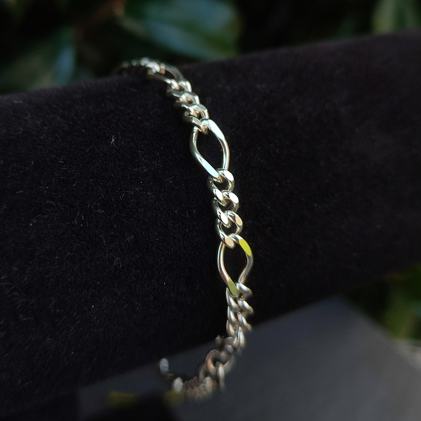 The silverhollowjewellery Figaro Chain Bracelet in sterling silver is shown on a black velvet stand with a blurred green background.