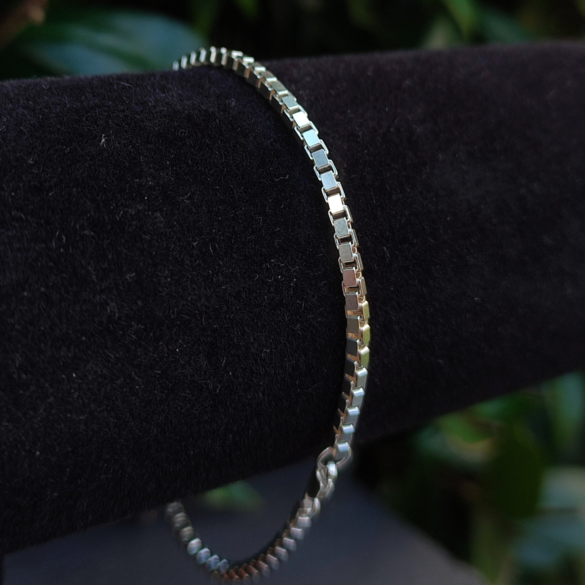 The Box Chain Bracelet- Sterling Silver by silverhollowjewellery is shown on a black velvet stand with a blurred green background.