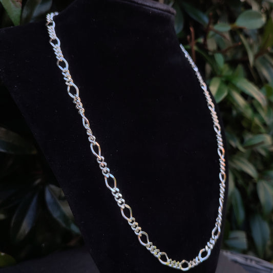 The silverhollowjewellery Figaro Chain Necklace in sterling silver features alternating diamond cut curb links, displayed on a black velvet stand with green foliage in the background.