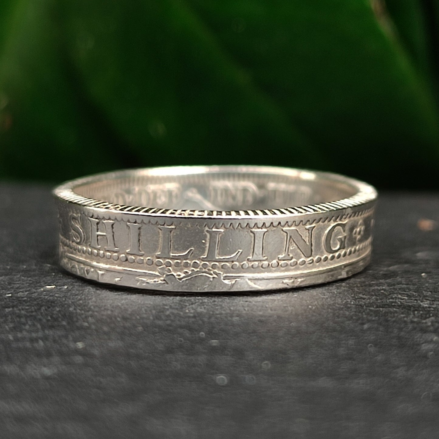 Handcrafted by silverhollowjewellery, the 1800s Victorian Shilling Ring in sterling silver features "SHILLING" engraved on an antique coin, showcased against a dark backdrop with green foliage.