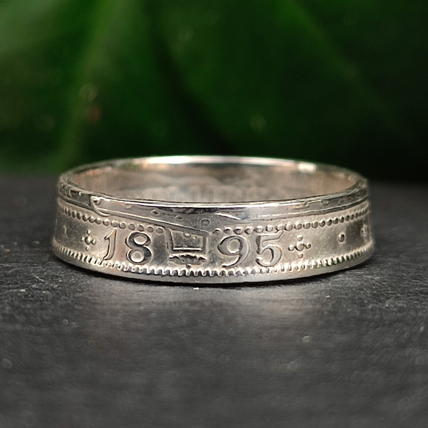 The silverhollowjewellery 1800s Victorian Shilling Ring, crafted from sterling silver and engraved with "1895" and ornate patterns, is displayed on a dark surface with a blurred green background.