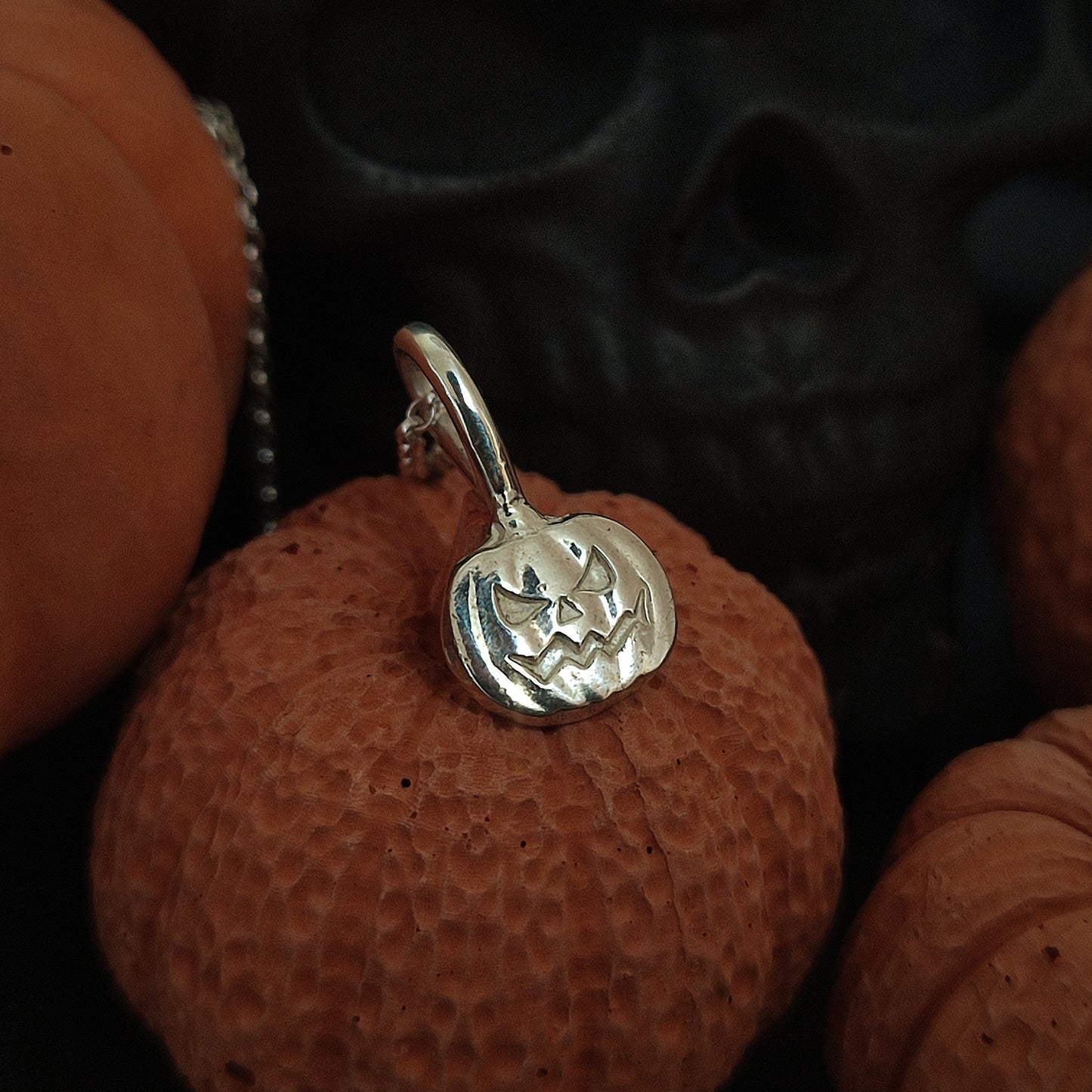 The Pumpkin Pendant- Sterling Silver by silverhollowjewellery features a handcrafted carved face design, shown on a textured orange pumpkin with a dark skull in the background.