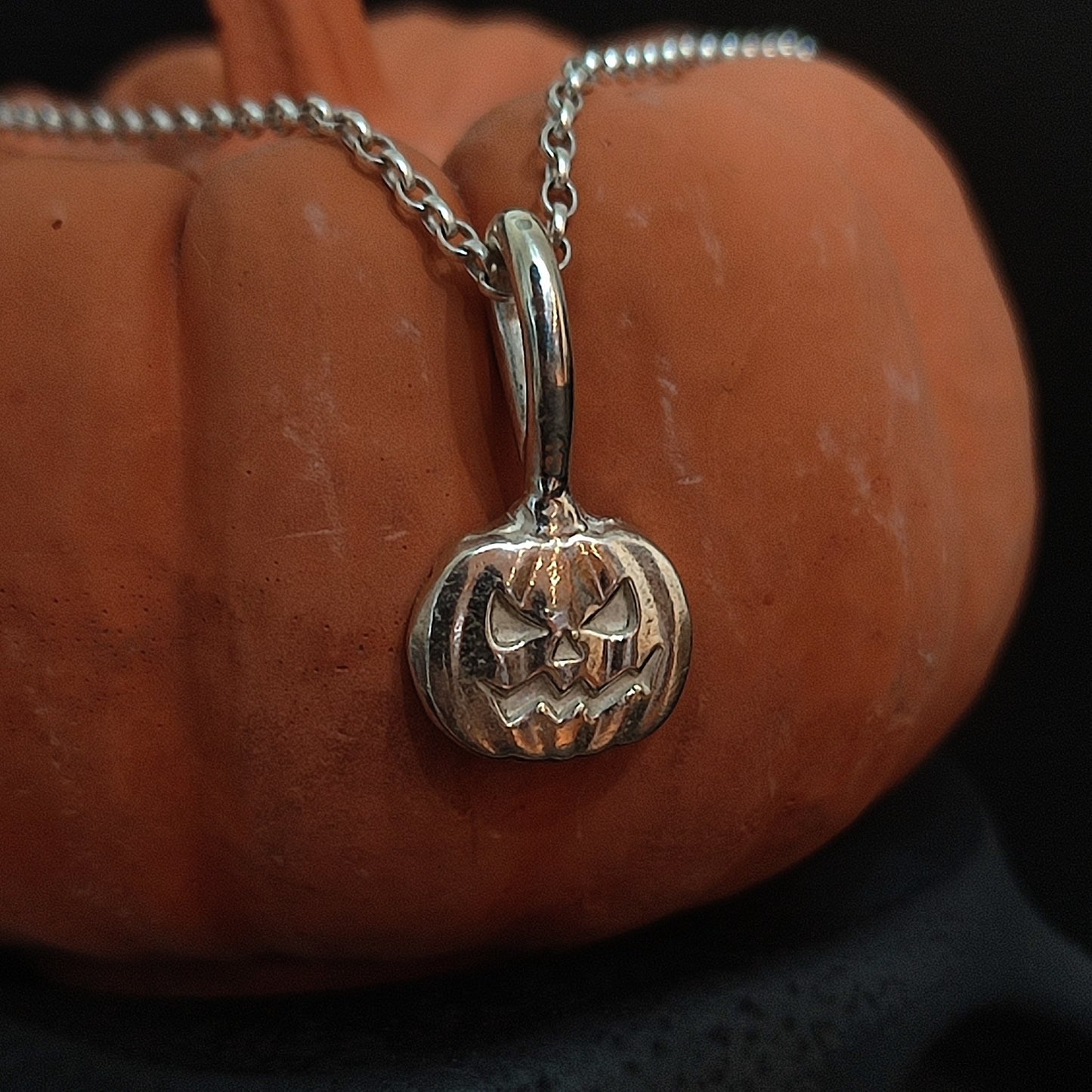 The silverhollowjewellery Pumpkin Pendant—crafted from sterling silver in a jack-o'-lantern shape—hangs on a chain and is displayed before an orange pumpkin.