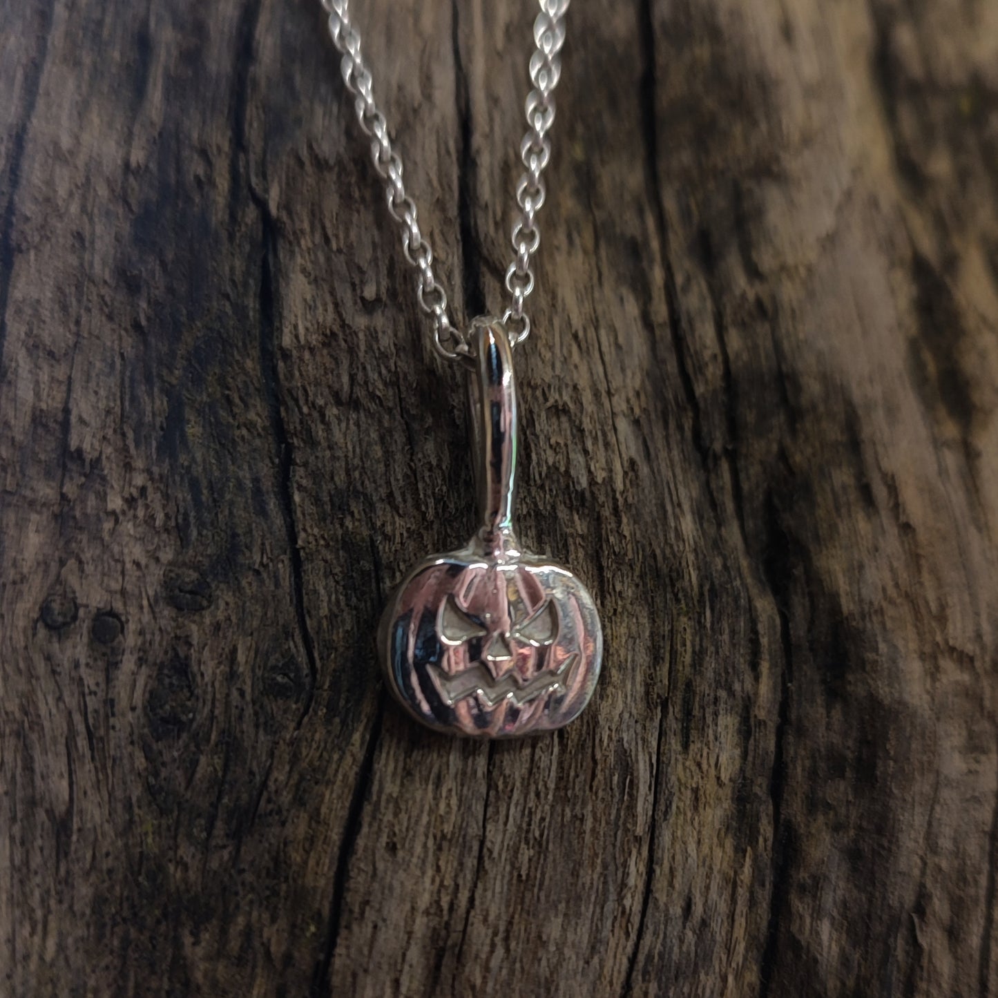 The Pumpkin Pendant–Sterling Silver by silverhollowjewellery, a handcrafted necklace featuring a jack-o’-lantern design, rests on a textured wooden surface.
