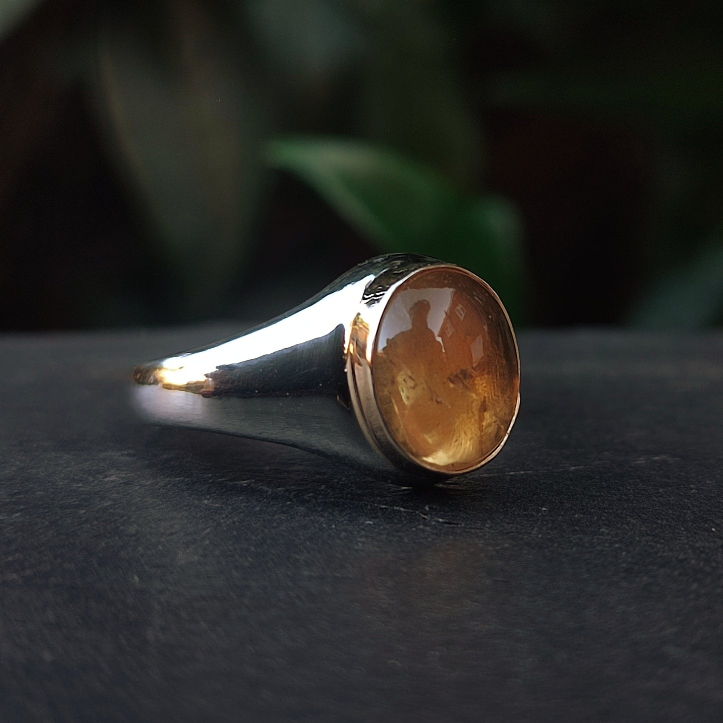 The Gold Bezel Citrine Ring by silverhollowjewellery features a polished yellow-brown citrine gemstone set in sterling silver, displayed on a dark surface with blurred green foliage in the background.