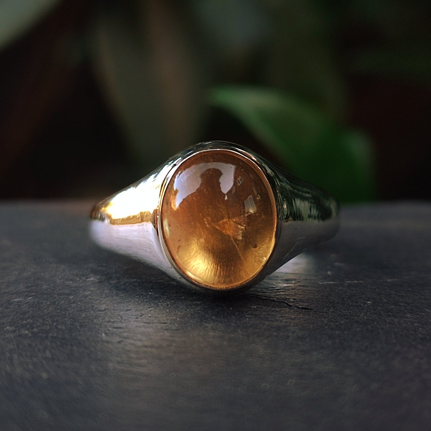 The Gold Bezel Citrine Ring by silverhollowjewellery is showcased with a smooth citrine stone set in gold, displayed on a dark surface against a blurred background.