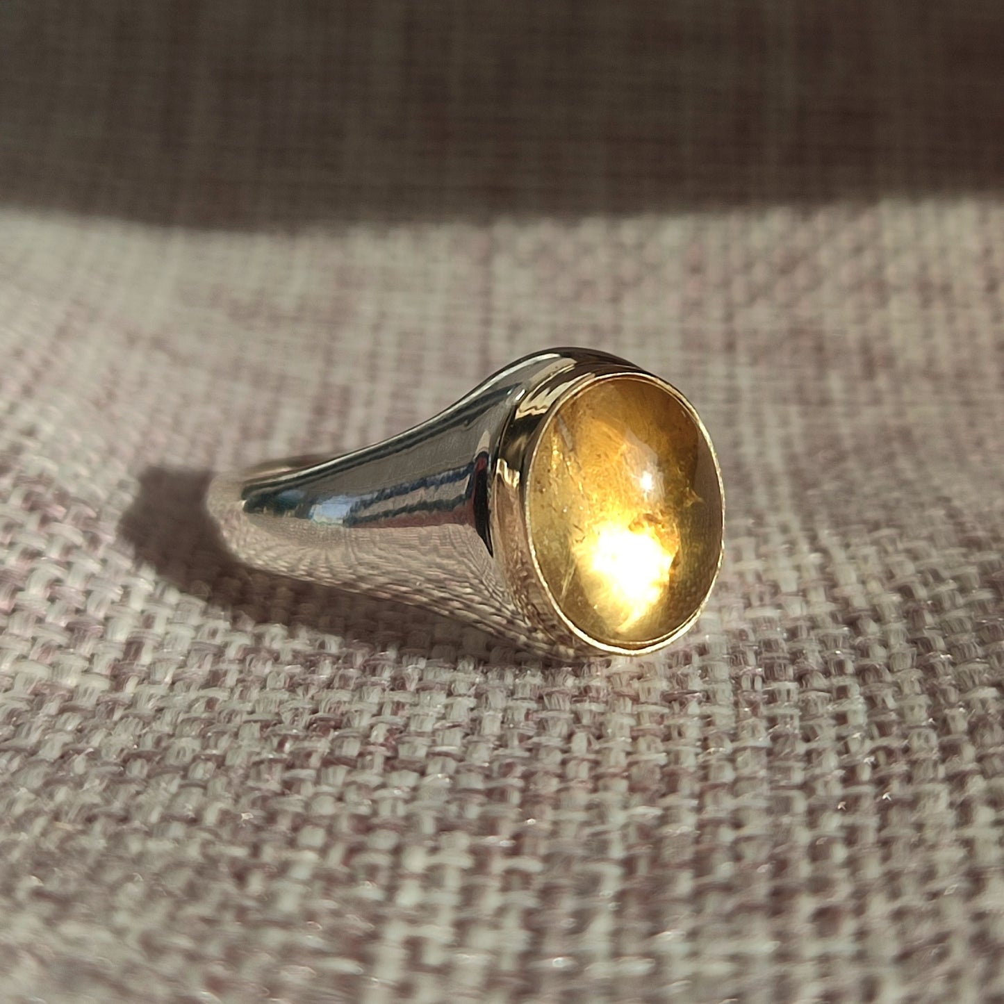 The Gold Bezel Citrine Ring by silverhollowjewellery features a polished citrine gemstone set in a smooth gold bezel, displayed on textured pink and white fabric in sunlight.