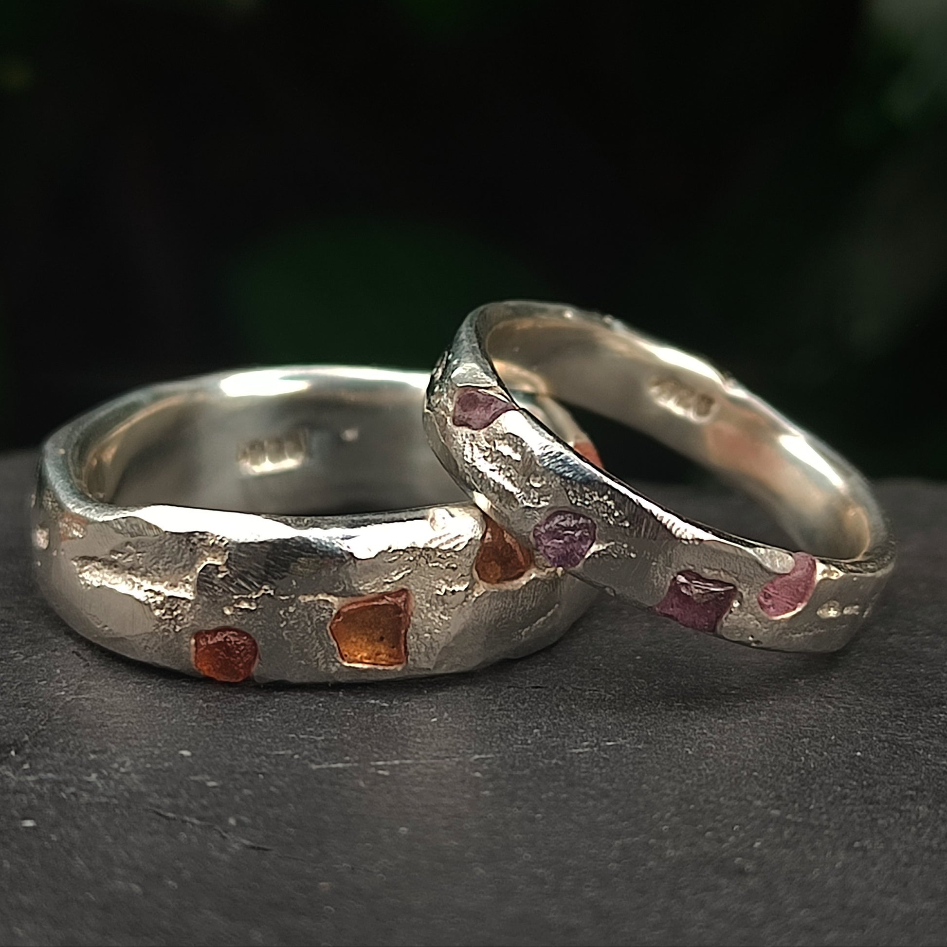 Two Raw Sapphire Band Sterling Silver rings by silverhollowjewellery feature hammered textures and raw sapphires in vibrant orange and purple, displayed on a dark surface.
