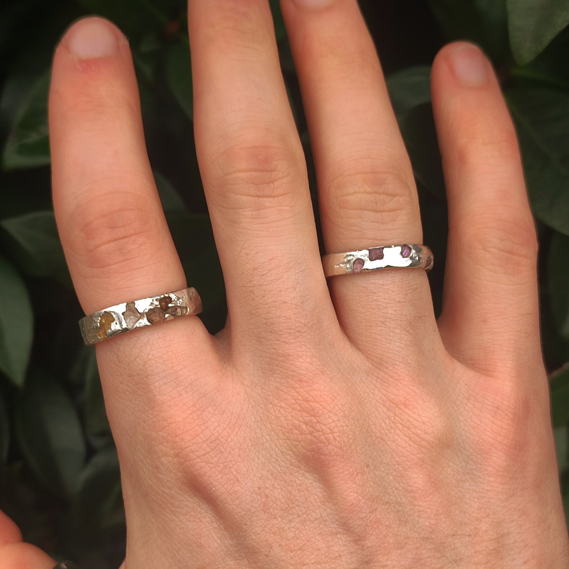 A hand wearing two silverhollowjewellery Raw Sapphire Band Sterling Silver - Custom rings on the middle and ring fingers, set against a backdrop of green leaves.