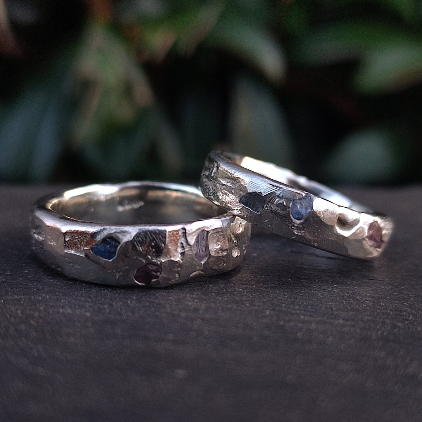 Two Raw Sapphire Band Sterling Silver rings by silverhollowjewellery, crafted from recycled sterling silver with a hammered finish and raw sapphire gemstones, are displayed on a dark surface with blurred greenery in the background.