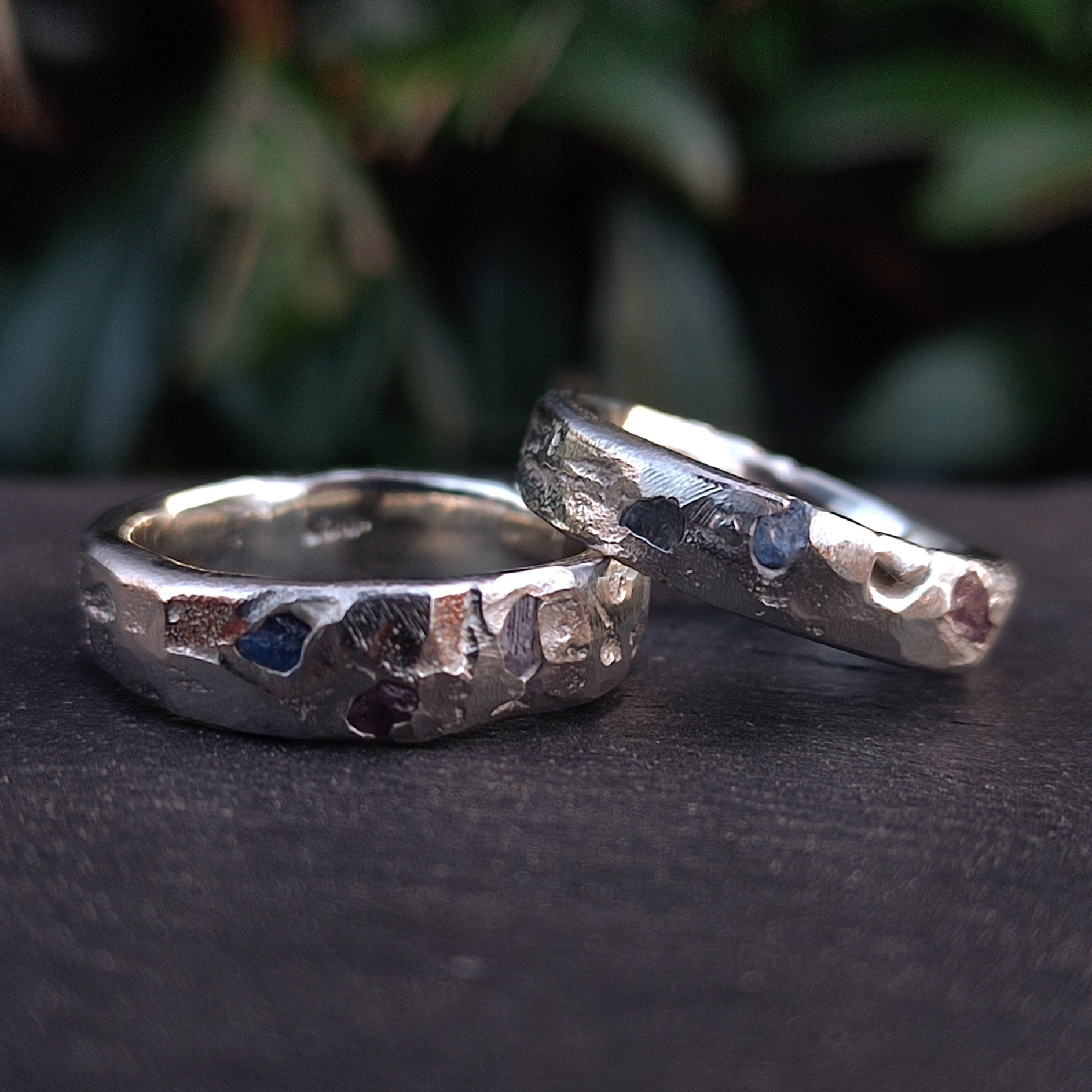 Two Raw Sapphire Band Sterling Silver rings by silverhollowjewellery, crafted from recycled sterling silver with a hammered finish and raw sapphire gemstones, are displayed on a dark surface with blurred greenery in the background.