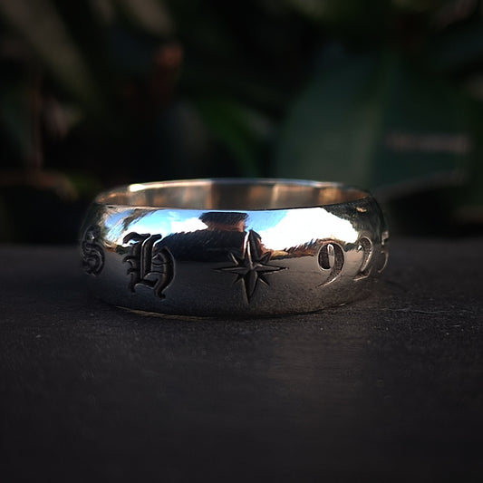 The Silver Makers Mark Ring by silverhollowjewellery, a chunky sterling silver piece featuring engraved letters, a star symbol, and the number 922, sits on a dark surface with green foliage in the background.