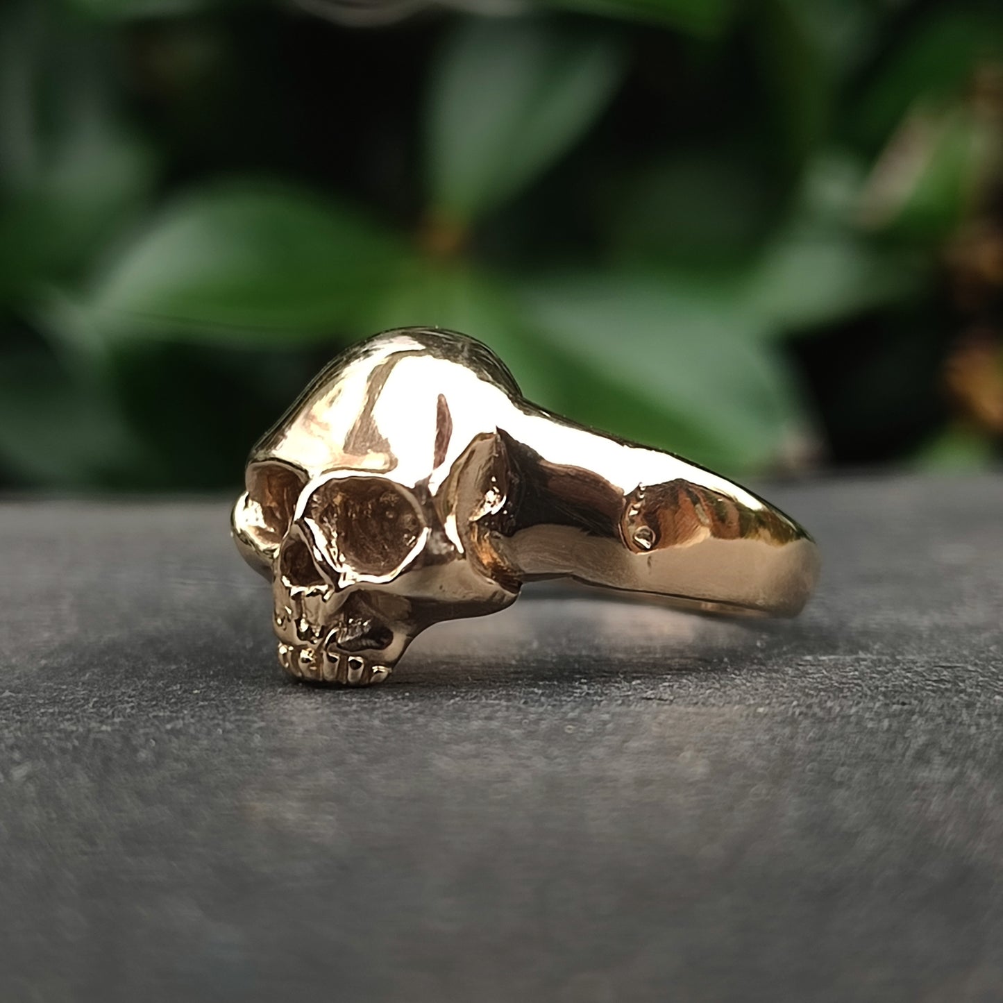 The Skull Ring 9ct Gold by silverhollowjewellery is a handcrafted yellow gold ring with a detailed skull design, shown resting on a gray surface with green foliage softly blurred in the background.
