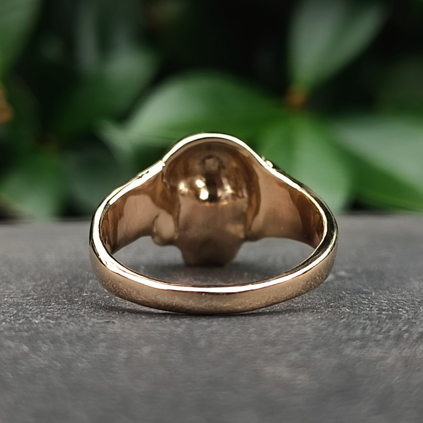 Close-up of the Skull Ring 9ct Gold by silverhollowjewellery, handcrafted in yellow gold and displayed on a gray surface with blurred green leaves in the background.