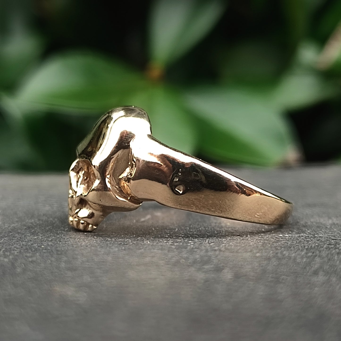The silverhollowjewellery Skull Ring 9ct Gold features a handcrafted sculpted skull design, shown against a backdrop of blurred green foliage.
