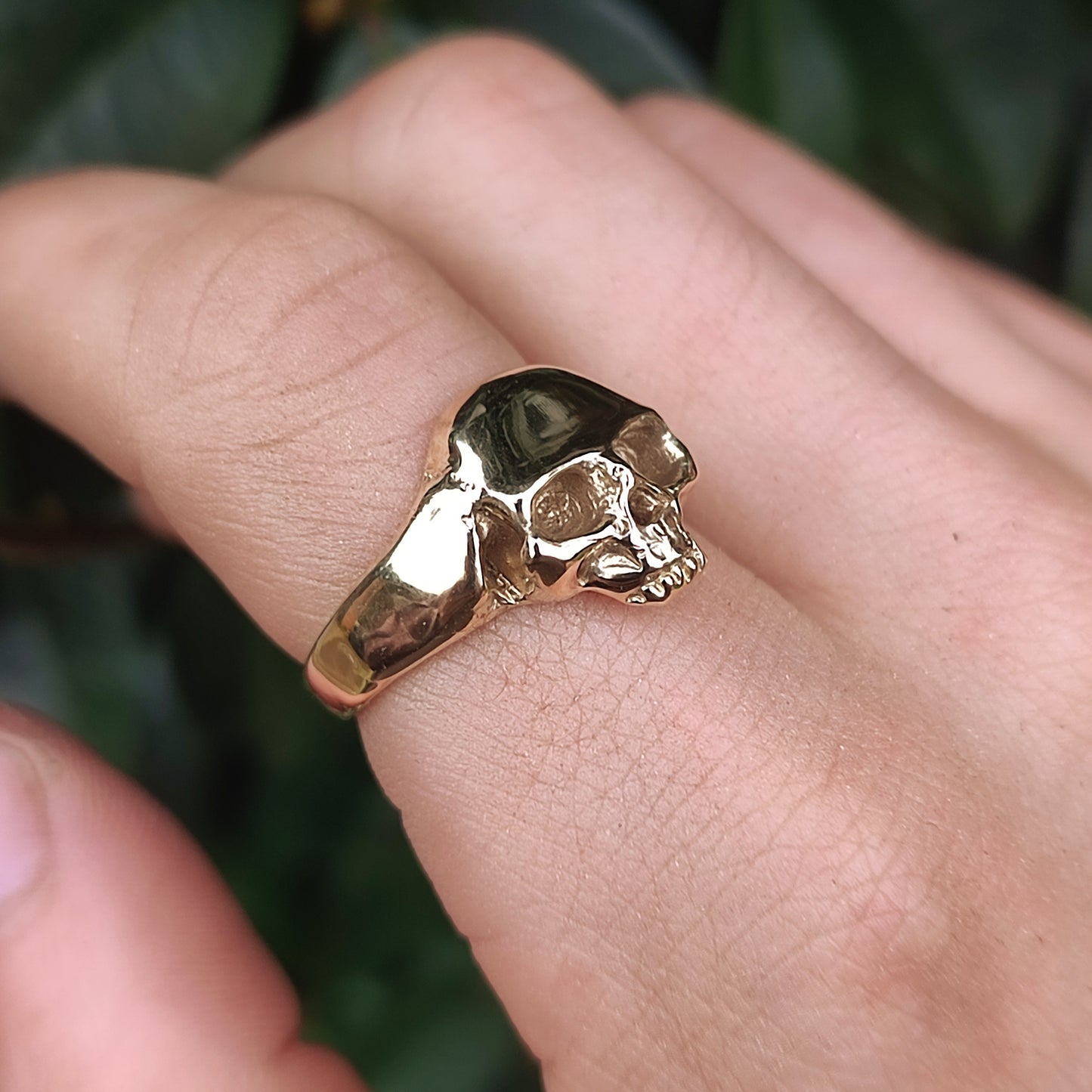 The Skull Ring 9ct Gold by silverhollowjewellery is worn on a person's finger, set against a green leafy background.