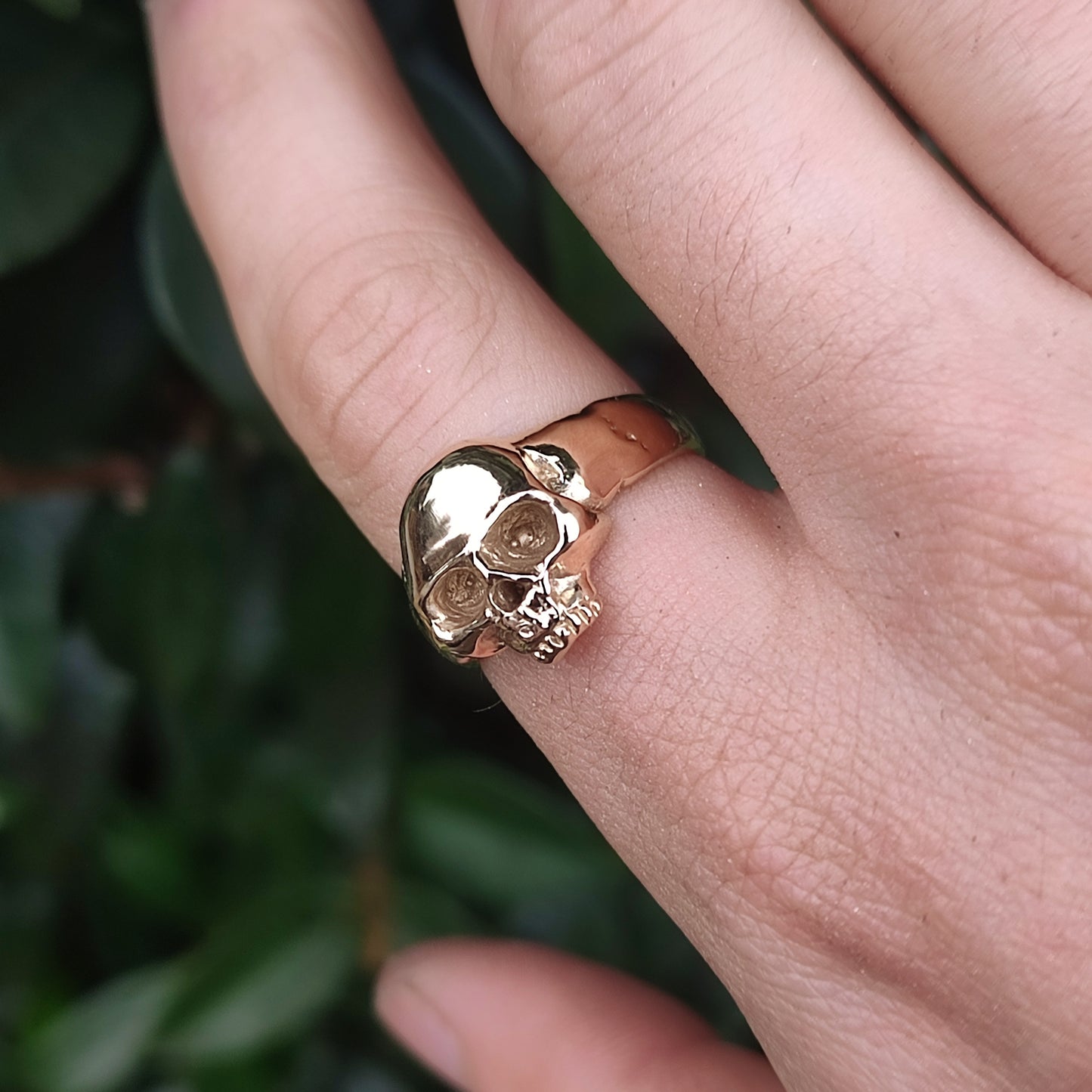 A close-up of a hand wearing the silverhollowjewellery Skull Ring 9ct Gold, with softly blurred green leaves in the background.