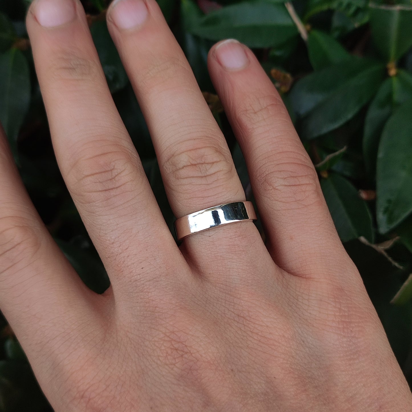 A hand wears the silverhollowjewellery Plain Band Sterling Silver ring, handcrafted from recycled 925 sterling silver, on the middle finger above green leaves.