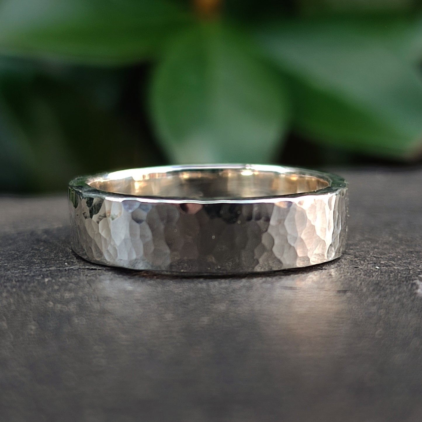 The Chunky Hammered Ring Sterling Silver by silverhollowjewellery, crafted from recycled 925 sterling silver, rests on a stone surface with soft green leaves blurred in the background.