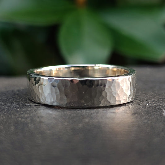 The Chunky Hammered Ring Sterling Silver by silverhollowjewellery, crafted from recycled 925 sterling silver, rests on a stone surface with soft green leaves blurred in the background.