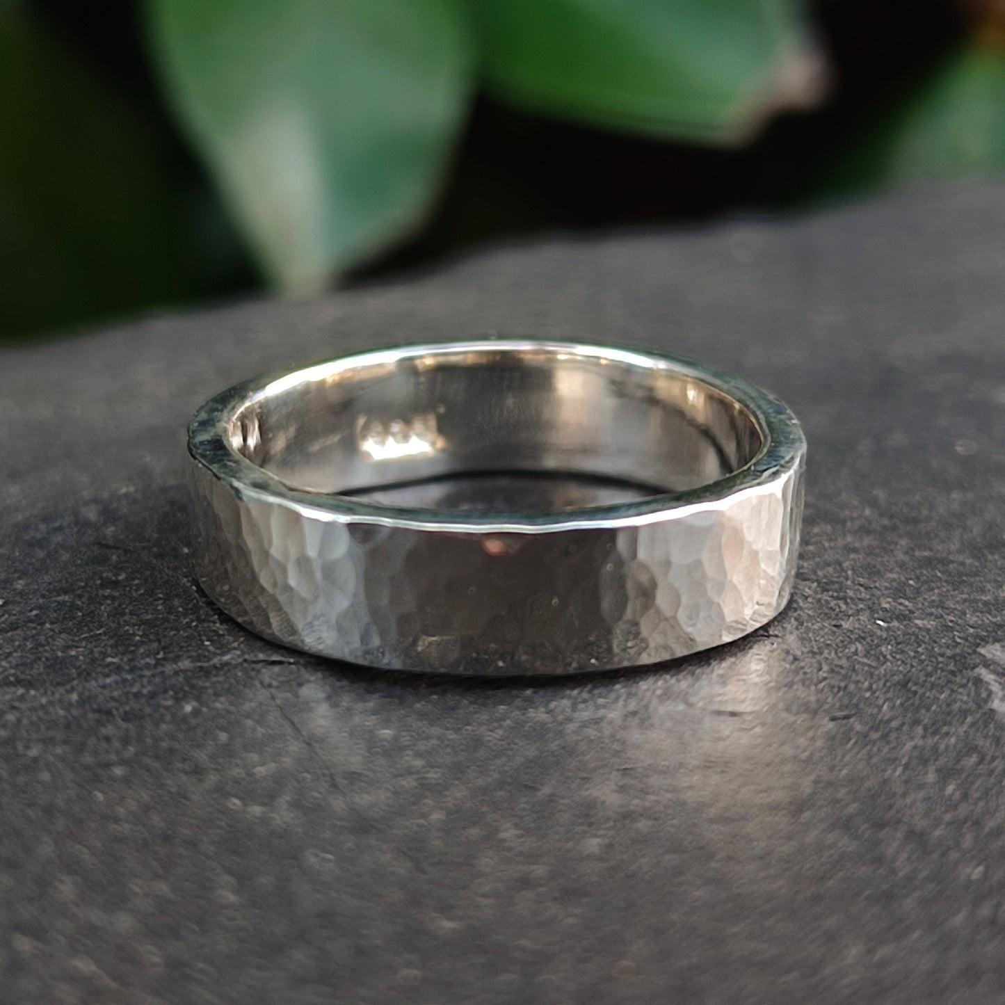 The Chunky Hammered Ring Sterling Silver by silverhollowjewellery, made from recycled 925 sterling silver, sits on a dark stone surface with blurred green leaves in the background.