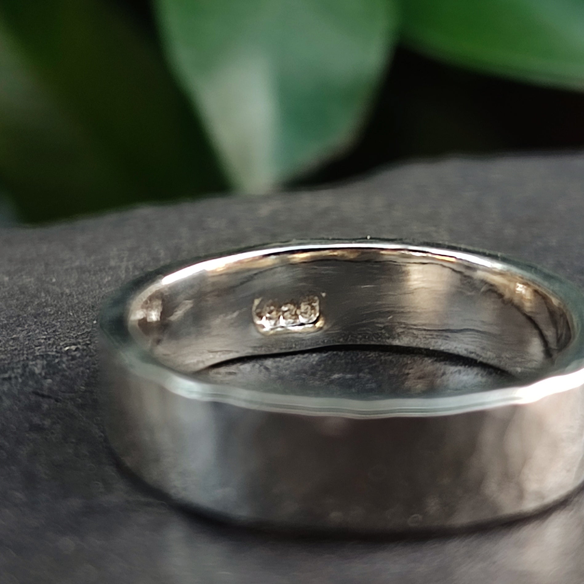 A close-up of the Chunky Hammered Ring Sterling Silver by silverhollowjewellery, crafted from recycled 925 sterling silver with a stamped hallmark on the inner band, rests on a dark surface with blurred green leaves in the background.
