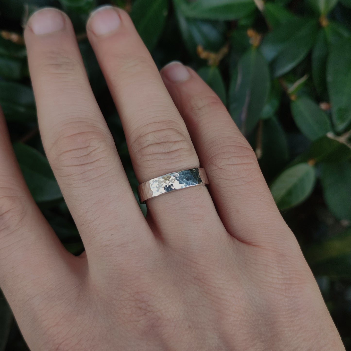 A hand wears the silverhollowjewellery Chunky Hammered Ring Sterling Silver on the middle finger, crafted from recycled 925 sterling silver, against green leaves.