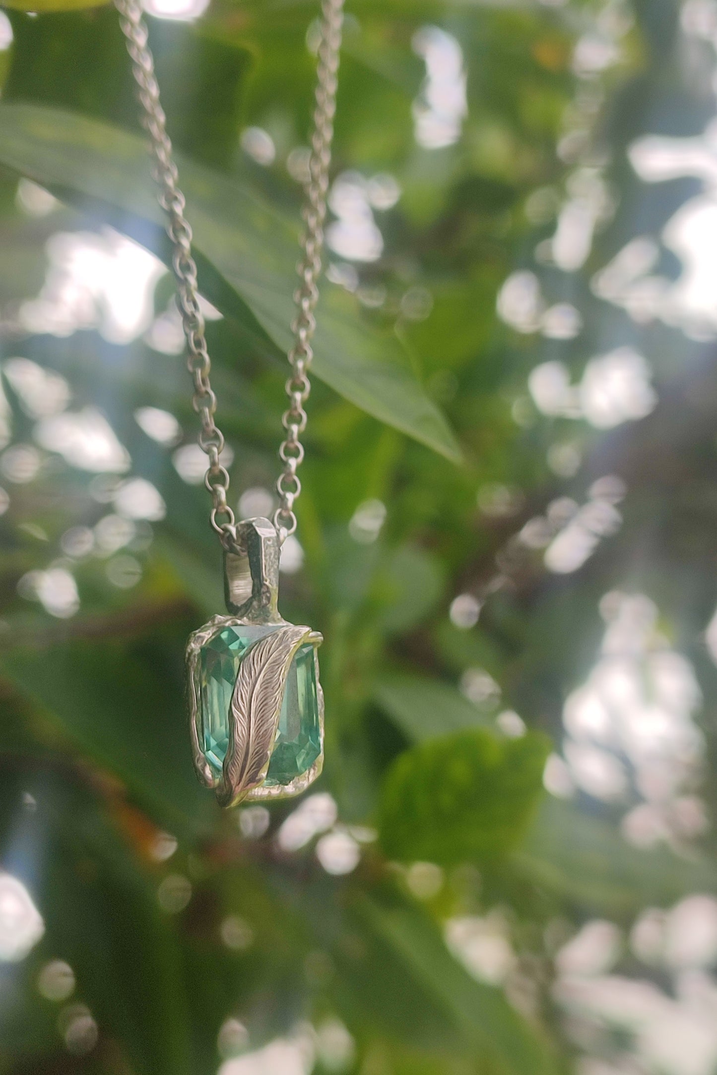 The Feather Teal Sapphire Pendant by silverhollowjewellery features a sterling silver chain and green gemstone in a feather setting, displayed outdoors with blurred green foliage.