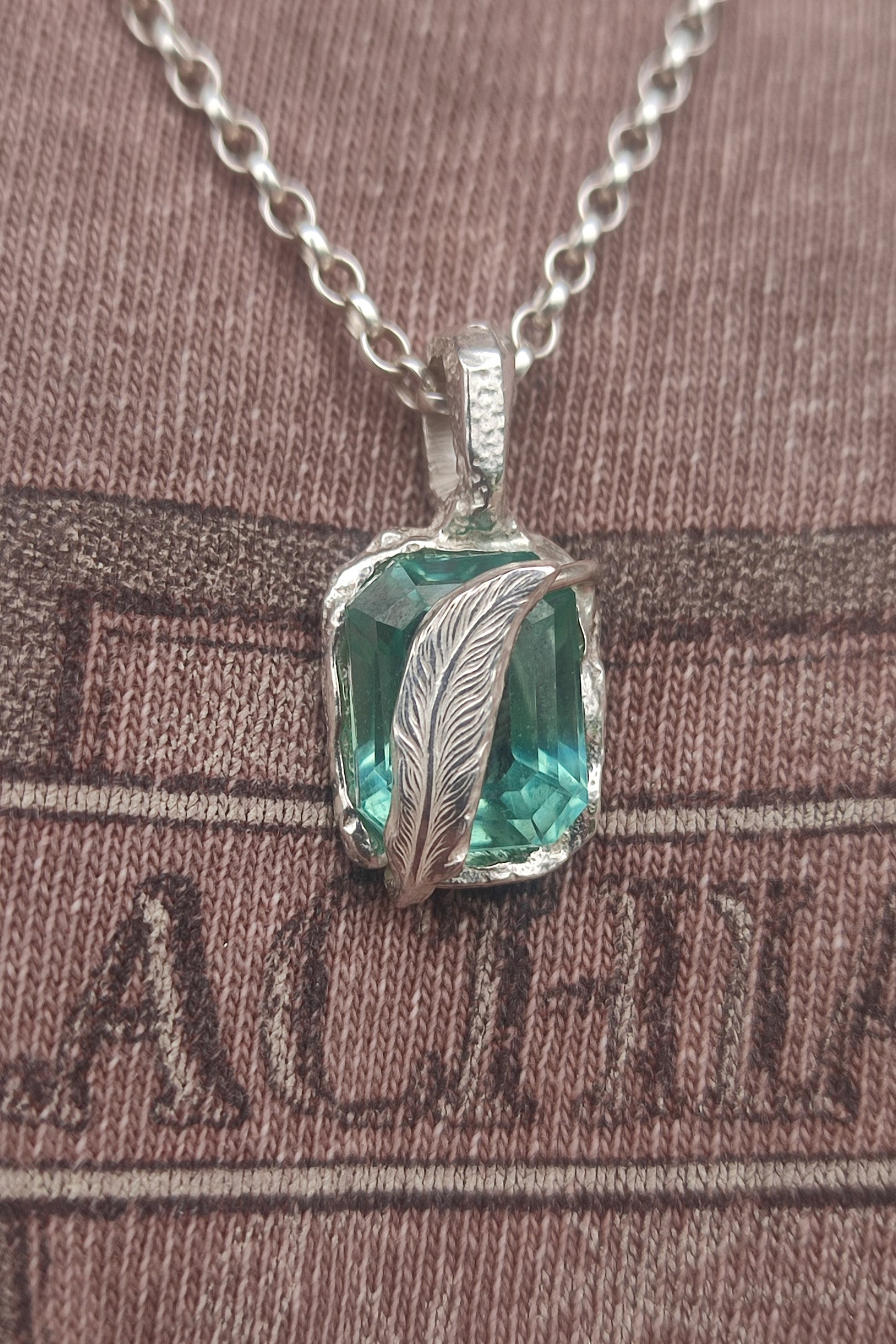 The Feather Teal Sapphire Pendant by silverhollowjewellery features a square teal sapphire in a detailed sterling silver feather setting, presented on a sterling silver chain against a brown textured fabric background.