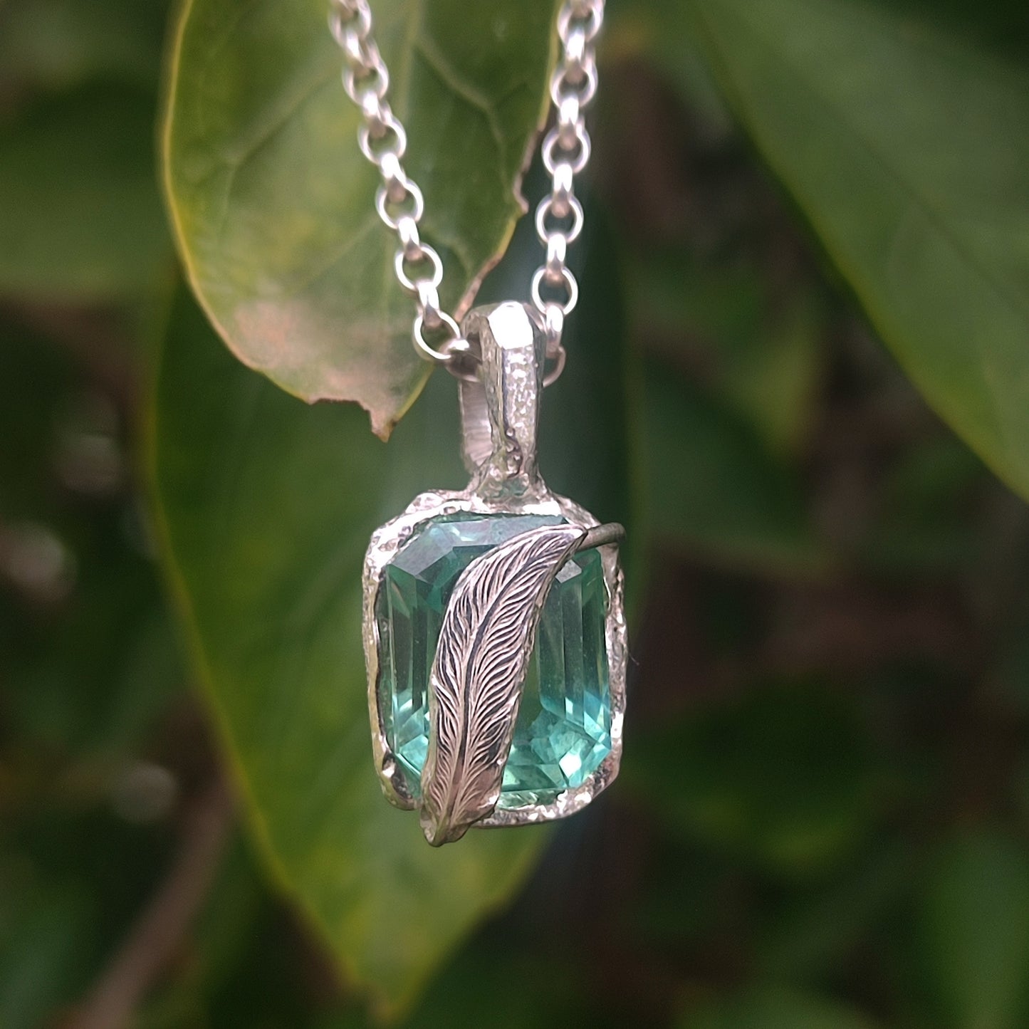 The Feather Teal Sapphire Pendant in sterling silver by silverhollowjewellery features a delicate feather setting, showcased against green leaves.