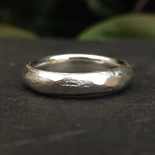 A close-up of the Raw Band by silverhollowjewellery—an organic, textured ring crafted from recycled 925 sterling silver with a unique uneven surface—set against a dark background with blurred green foliage.