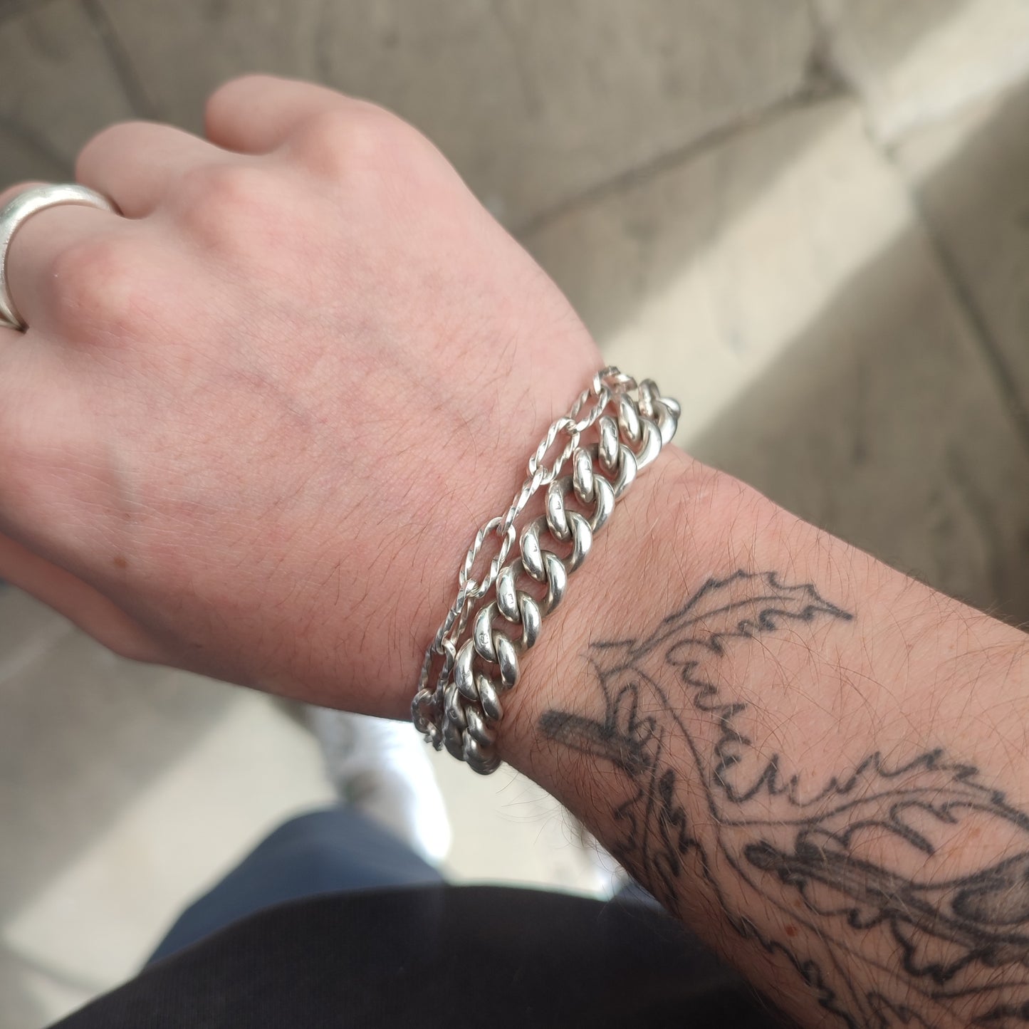 A wrist adorned with silverhollowjewellery’s Silver Watch Chain Bracelet, a silver ring on the middle finger, and a leafy branch tattoo winding along the forearm.
