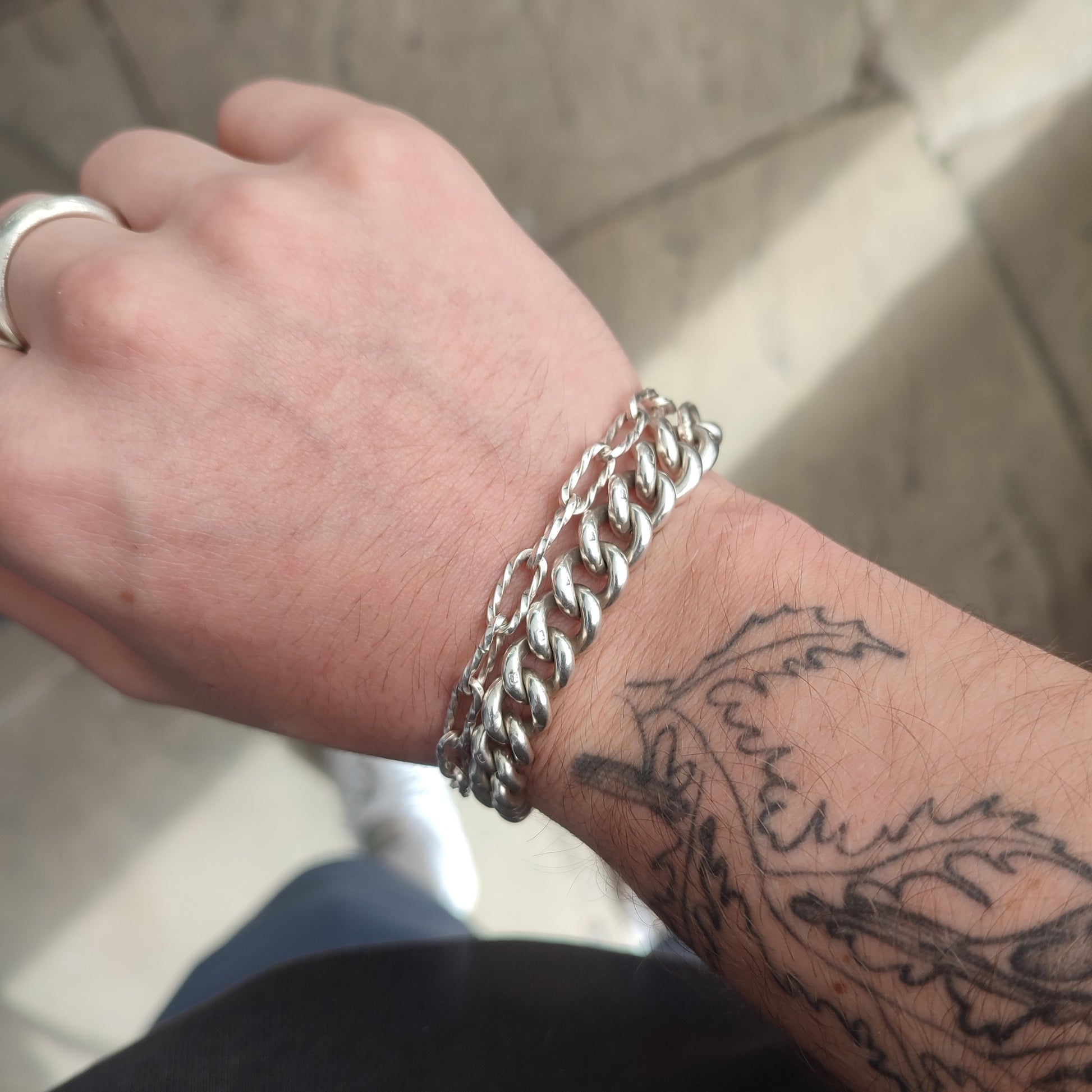 A wrist adorned with silverhollowjewellery’s Silver Watch Chain Bracelet, a silver ring on the middle finger, and a leafy branch tattoo winding along the forearm.