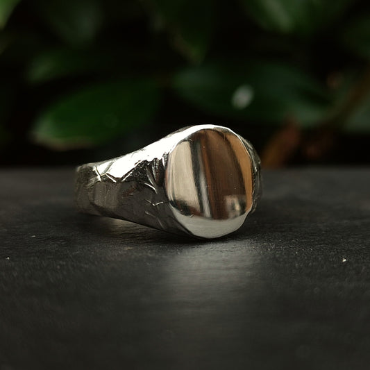The Oval Rock Signet by silverhollowjewellery, a handcrafted sterling silver ring with a smooth, blank oval face, rests on a dark surface with blurred green leaves in the background.
