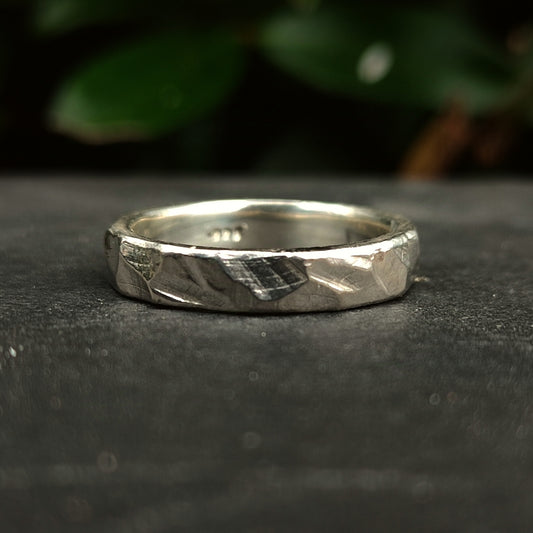 The Thin Rock Band by silverhollowjewellery—a handcrafted, rock-textured ring made from recycled 925 sterling silver—rests on a dark, smooth surface with blurred green leaves in the background.