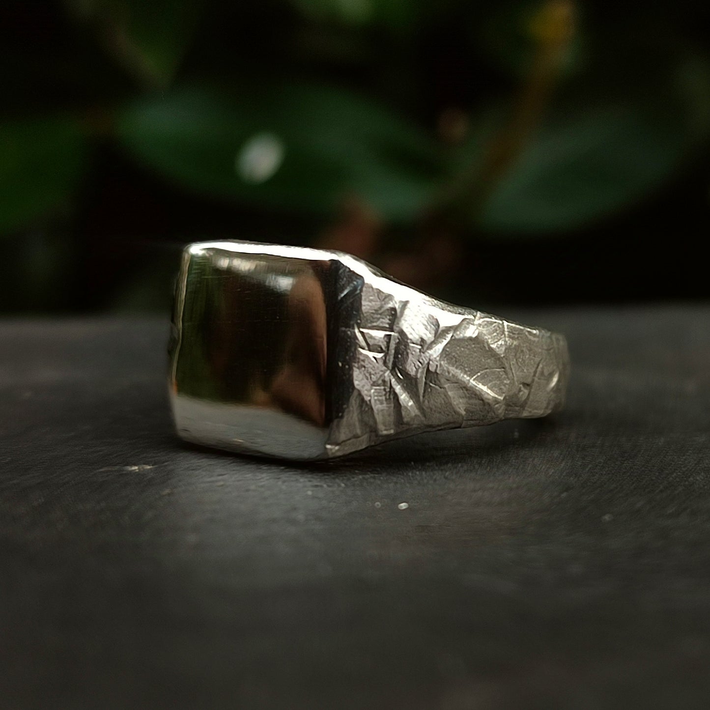The Square Rock Signet by silverhollowjewellery, crafted from recycled sterling silver, features a rock-textured band and a smooth rectangular face, shown resting on a dark surface with blurred green foliage behind.