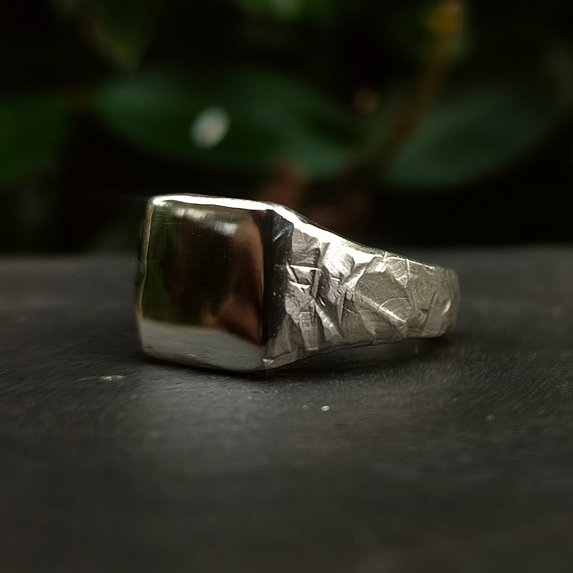 The Square Rock Signet by silverhollowjewellery, crafted from recycled sterling silver, features a rock-textured band and a smooth rectangular face, shown resting on a dark surface with blurred green foliage behind.