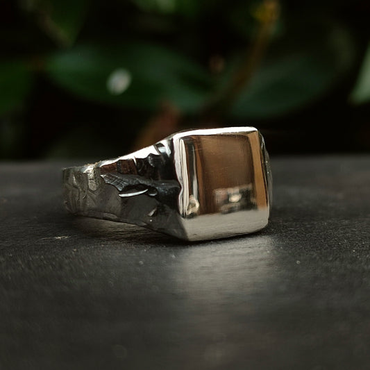 The Square Rock Signet by silverhollowjewellery, crafted from recycled sterling silver, features a smooth rectangular face and a rock-textured band, displayed on a dark surface with blurred greenery in the background.