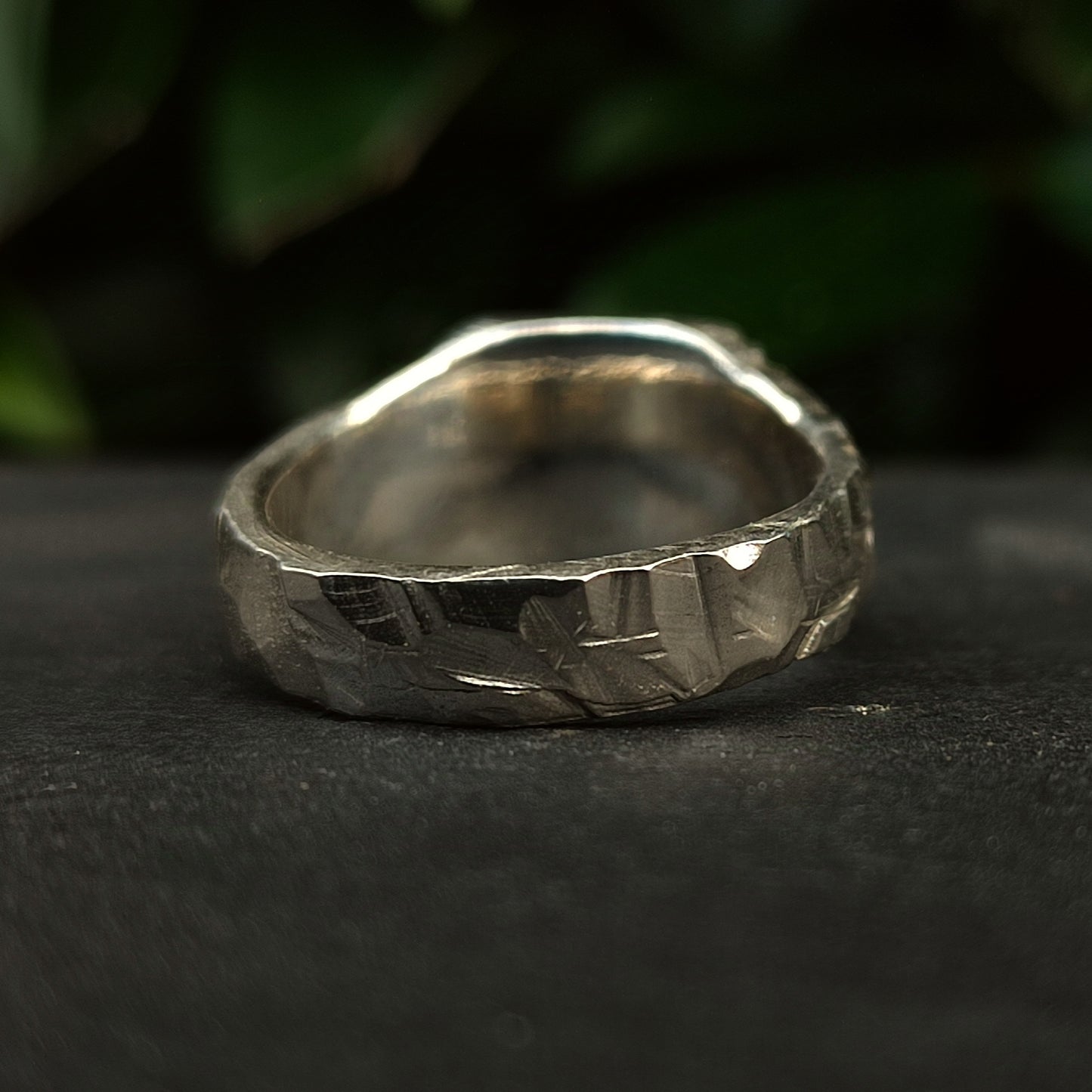 The Square Rock Signet by silverhollowjewellery features a rock-textured, hammered finish in recycled sterling silver. It rests on a dark surface with blurred green foliage in the background.