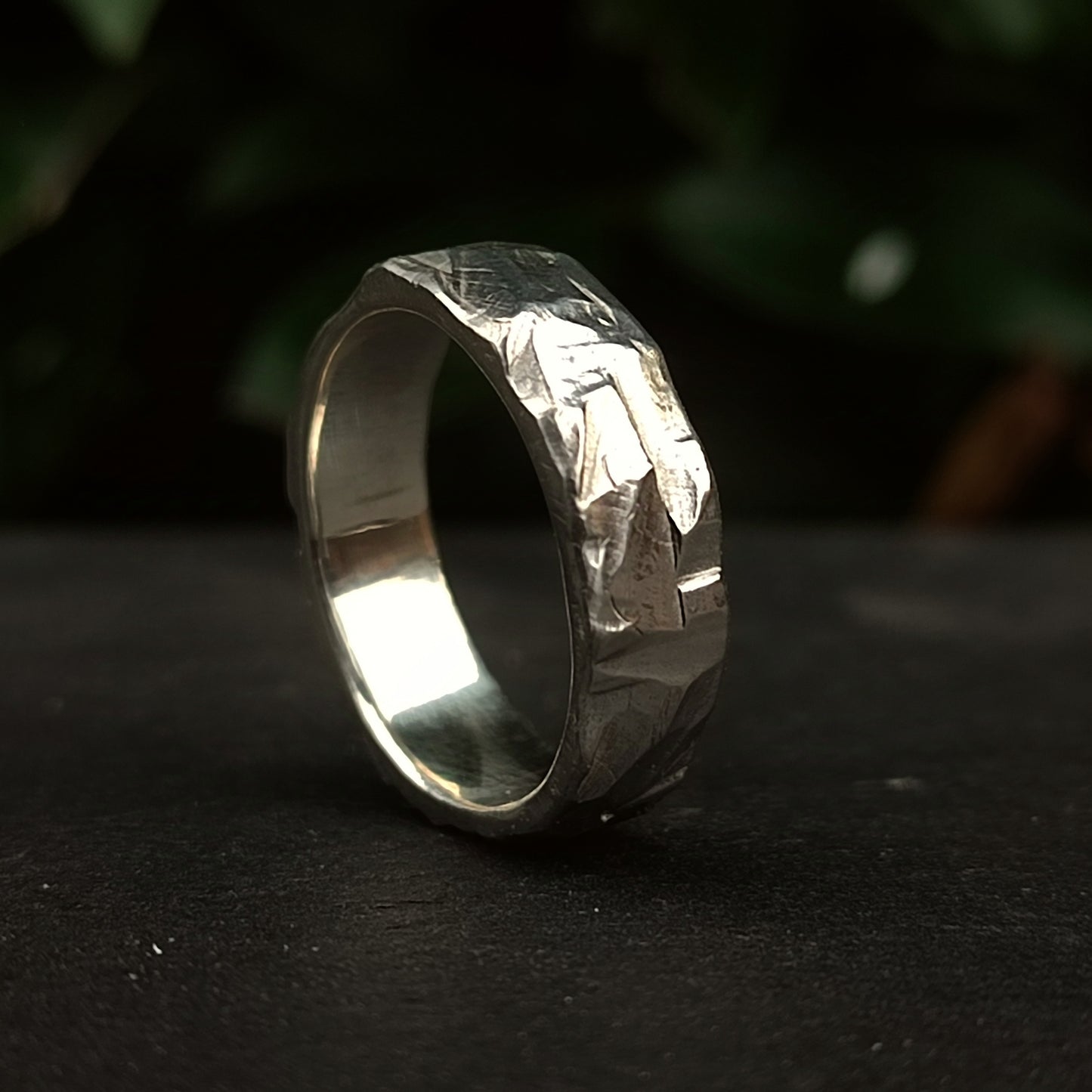 The Thick Rock Band by silverhollowjewellery, made from recycled silver with a rock-textured, hammered finish, stands upright on a dark surface against a blurred green background.